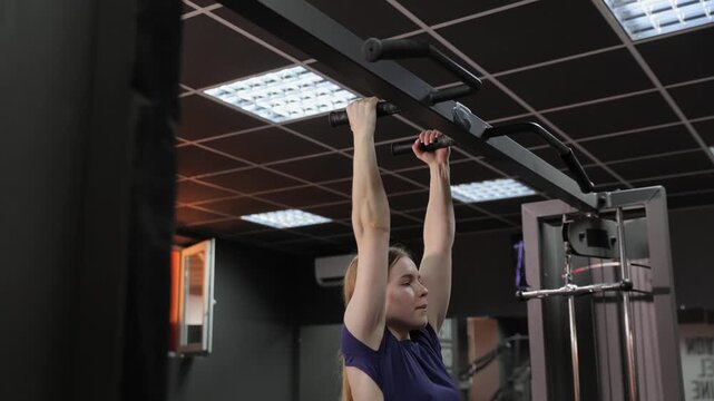 Dedicated fit woman strengthening her back and biceps muscles while performing pull ups on a horizontal bar during an intense workout session inside a modern and dark gym