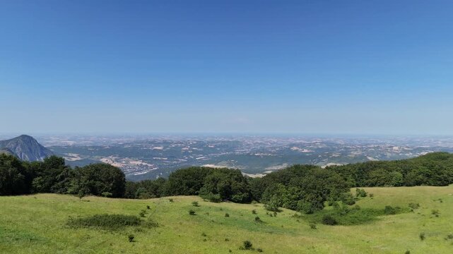 Vista panoramica da un'altura verde su una vasta vallata pianeggiante, con colline boscose in primo piano e cielo azzurro terso all'orizzonte: scorcio suggestivo di paesaggio montano italiano.