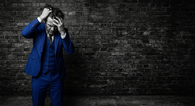 Man in blue suit holding head in distress against dark textured wall copy space
