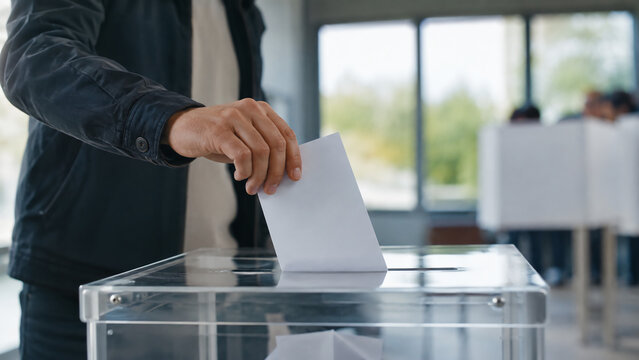 A person deposits a ballot into a transparent voting box in a bright room with voters and booths in the background.