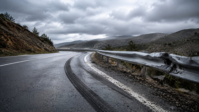 A winding mountain road with fresh tire skid marks under a cloudy sky, featuring guardrails and rugged terrain on both sides.
