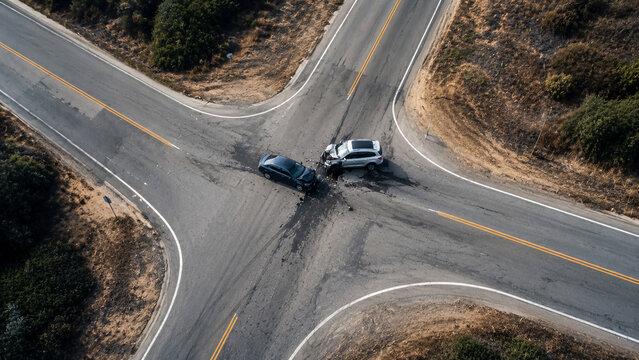 Two cars collided at a rural four-way intersection, causing visible damage and debris on the asphalt surrounded by dry vegetation.