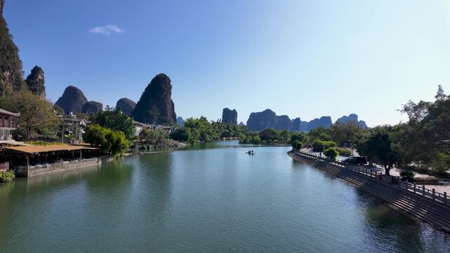 A calm river meanders past karst peaks, with lush trees and simple structures under a clear blue sky on a sunny day.