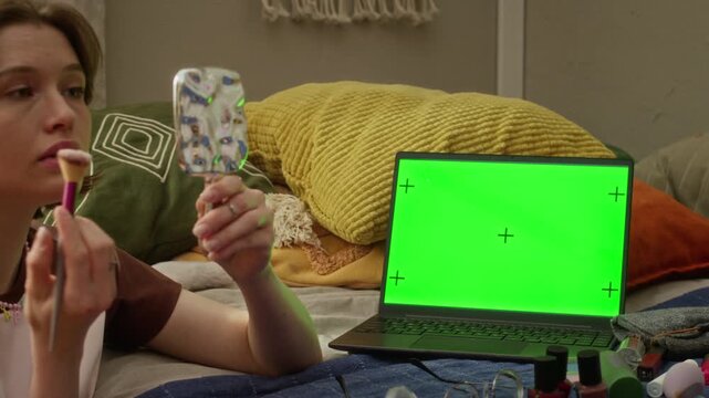 Young Caucasian woman applying makeup with cosmetic brush while looking in hand mirror next to green screen laptop. Clip is ideal for beauty vlogging, online tutorials, and tech integration