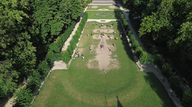 Aerial drone footage moving from top to bottom, revealing the historical stone Beyaert Tower (Tour de Tournai) and people relaxing on the green lawn in the Cinquantenaire Park, Brussels.