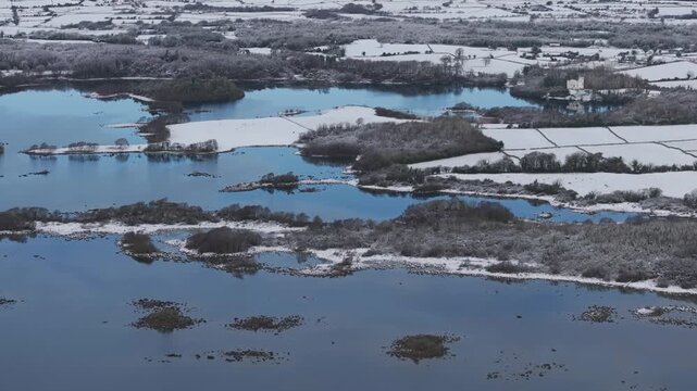 A cinematic aerial shot of the icy blue waters of Lough Corrib surrounded by snowcovered fields and frozen trees on a quiet winter morning in Ireland.