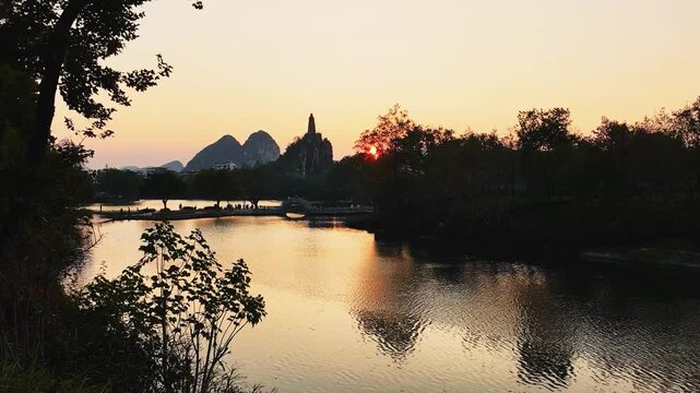 A tranquil landscape features a river reflecting the warm glow of sunset, with a pagoda and mountains in the distance.
