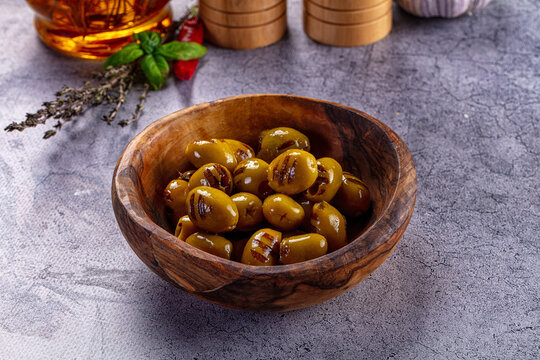 Grilled green olives in rustic wooden bowl with herbs and olive oil