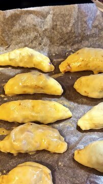 Rows of uncooked empanadas arranged on parchment paper are brushed with egg wash before baking in a kitchen setting.