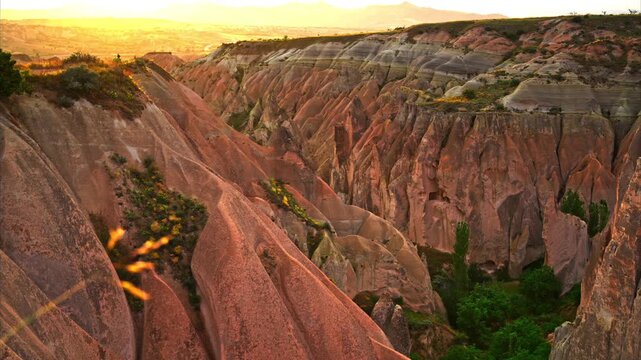 A high-angle shot of the unique geological formations in Cappadocia, Turkey, featuring layered volcanic tuff rocks, traditional fairy chimneys, and green vegetation in the valley. Sunset