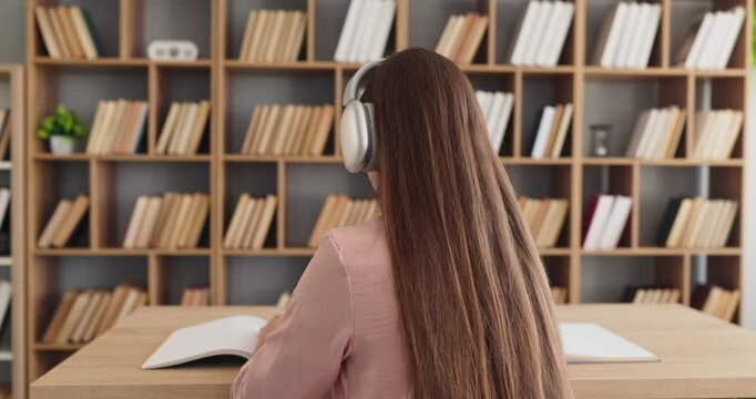 Student woman studying with headphones in library. Books and education drive concentration at a wooden desk. Rear view shows focused learning in a quiet room. Clear study focus concept image.