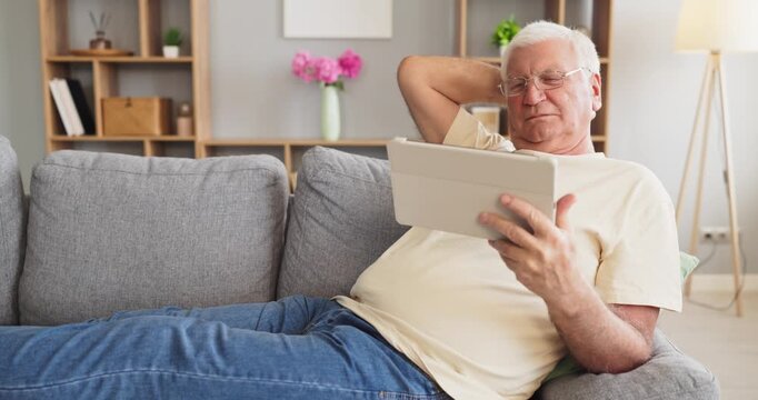 Senior elderly man online browsing tablet at home. Resting on a sofa, he reads, shops and chats, using technology for connection and leisure in retirement. Theme is senior tech lifestyle.