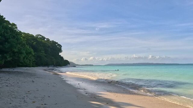 A panoramic view of a serene tropical beach featuring soft white sand, calm turquoise waters, and a lush green jungle on the shoreline under a bright blue sky with light clouds.