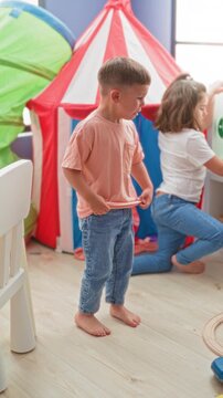 Boy standing barefoot with hands at waistband and girl kneeling by a red play tent in a building; childhood tension.