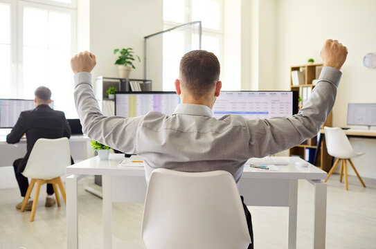Businessman employee celebrates success at office computer. Rear view man raises arms in victory after checking a spreadsheet on monitors in a bright workplace. Clear concept of finance success.