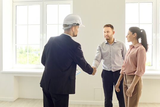 Couple and realtor handshake confirm apartment purchase. They seal agreement in a bright empty room, outline steps, plan renovation and review mortgage terms. Real estate trust and agreement.