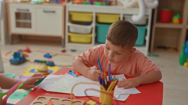 Boy drawing with crayons and colored pencils, hand holding blue pencil at red table in childcare building; creativity concentration.