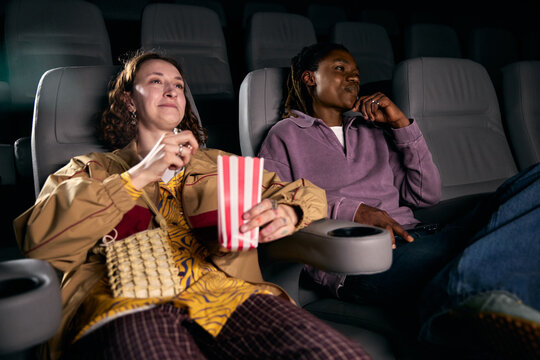 Caucasian woman and African American man watching movie, eating popcorn, sharing leisure time in cinema. Ideal for entertainment marketing, dating, diversity, lifestyle campaigns