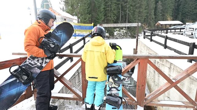 A man and child, equipped with snowboards, stand on a wooden bridge overlooking a small stream. The snowy landscape and wintery atmosphere create a peaceful scene.