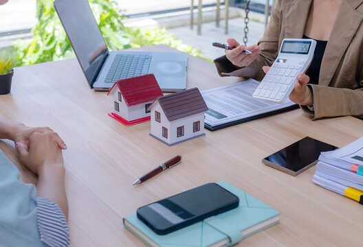 Real estate agent and a female client holding house models while discussing and comparing property options at the office desk.