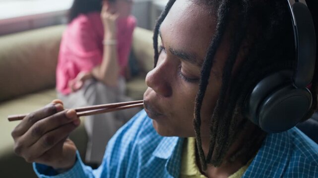 Tilt up of young adult Black man wearing headphones eating takeout food with chopsticks at office desk near laptop. Footage represents modern corporate lunch break and food delivery services