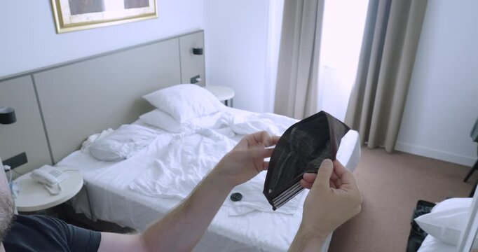 Man holding an empty wallet in front of a hotel bed after running out of money. Travel concept of financial problems, hotel expenses, budgeting, and accommodation costs.