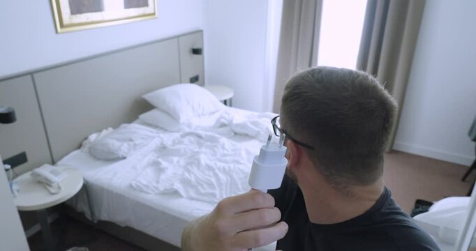 Man holding a power adapter and pointing at it in a hotel room, highlighting the problem of not having enough electrical outlets. Concept of travel, charging devices, and accommodation convenience.
