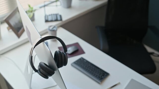 No people closeup shot of black wireless headphones hanging on computer monitor at empty office desk, representing hybrid work setup. Ideal for customer support, call center, remote business marketing