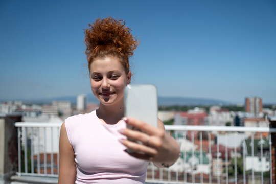 Beautiful and young redhead woman taking a selfie with a mobile phone on the rooftop of the city building