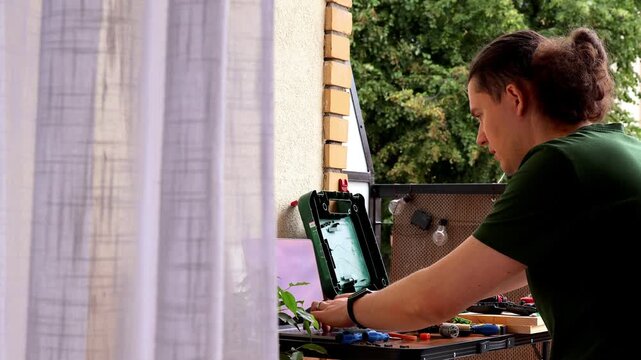 Man repairs cordless drill on balcony worktable while following online tutorial on laptop surrounded by tools and screwdrivers. Concept of right to repair, DIY maintenance, practical learning