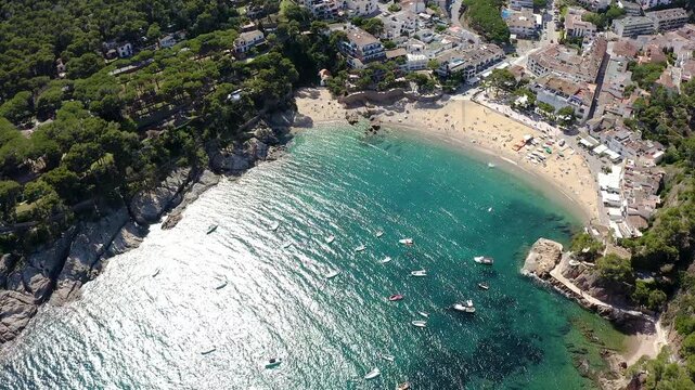 Aerial view of Tamariu Beach, a picturesque Mediterranean bay surrounded by pine-covered hills, golden sand, turquoise waters and the charming seaside atmosphere of the Costa Brava. 
