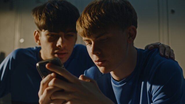 Young Soccer Players in Uniforms in Dressing Room, Sharing Content on Smartphone. One Boy Was Browsing Phone When Teammate Joined For Friendly Chat. Boys Smiling and Discussing Something on Screen.
