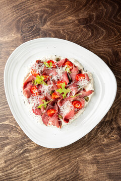 Beef carpaccio served on a creamy white sauce with cherry tomatoes, capers, greens, and shredded parmesan cheese on a white plate over a dark wooden table background.