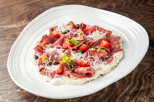 Beef carpaccio served on a creamy white sauce with cherry tomatoes, capers, greens, and shredded parmesan cheese on a white plate over a dark wooden table background.