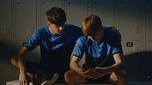 Two Young Soccer Players in Blue and Black Uniforms Sitting on Bench. One Boy Was Browsing Phone When Teammate Joined For Friendly Chat. Modern Youth Lifestyle, Digital Communication, and Friendship.