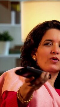 Indian Asian mother in her fifties with her pretty young daughter, both sitting on a cozy sofa at home, watching TV together with a remote in hand
