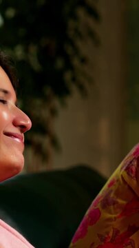 Indian Asian mother in her fifties with her pretty young daughter, both sitting on a cozy sofa at home, watching TV together with a remote in hand