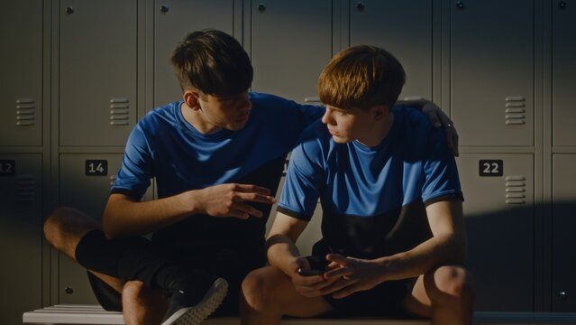 Two Young Soccer Players in Blue and Black Uniforms Sitting on Bench. One Boy Was Browsing Phone When Teammate Joined For Friendly Chat. Modern Youth Lifestyle, Digital Communication, and Friendship.