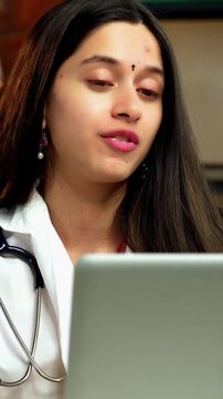 Indian Asian young female doctor in her mid-twenties, wearing a white coat, sitting at a desk with a laptop, conducting an online consultation with patients via video call in a professional setup