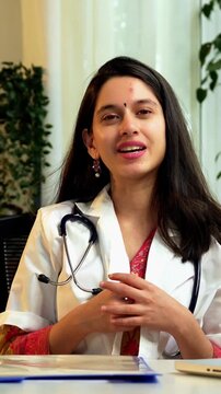 Indian Asian young female doctor in her mid-twenties wearing a coat, sitting at a desk with a laptop, smiling and looking at the camera while explaining something