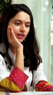 Indian Asian young female doctor in her mid-twenties, wearing a white coat, sitting at a desk with a laptop, conducting an online consultation with patients via video call in a professional setup