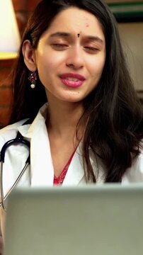Indian Asian young female doctor in her mid-twenties wearing a coat, sitting at a desk with a laptop, smiling and looking at the camera while explaining something