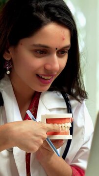 Indian Asian young female dentist wearing a white coat, sitting at a desk with a laptop, explaining oral health care using a model denture during a video call