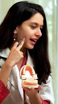 Indian Asian young female dentist wearing a white coat, sitting at a desk with a laptop, explaining oral health care using a model denture during a video call