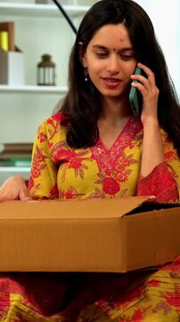Indian Asian angry young girl in her twenties wearing salwar kameez sitting on a sofa with a cardboard parcel, expressing frustration while talking to customer care over the phone about an issue