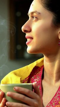 Indian Asian happy and pretty young girl in her twenties wearing salwar kameez and shawl, enjoying hot coffee or beverage in a modern kitchen with warm morning sunlight streaming in