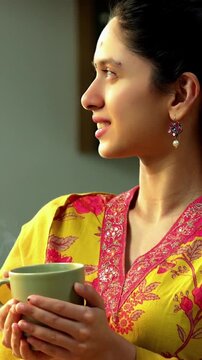 Indian Asian happy and pretty young girl in her twenties wearing salwar kameez and shawl, enjoying hot coffee or beverage in a modern kitchen with warm morning sunlight streaming in