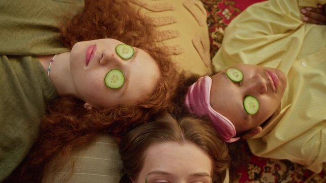 Diverse young adult women lying on floor during home spa day, wearing eye patches and cucumber slices on eyes with one eating slice. Ideal for beauty, wellness, or skincare marketing campaigns