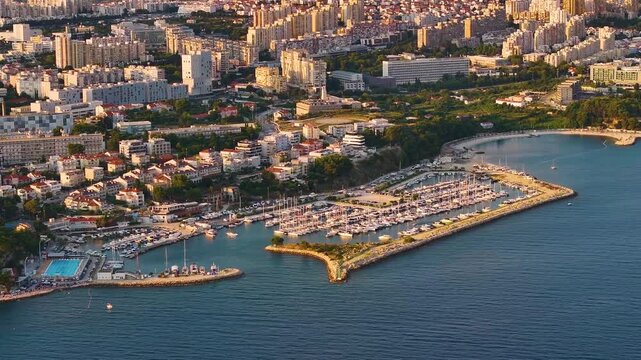 A view of the harbor in Split Croatia. Boats are docked in the marina. Buildings are visible in the background. The coastline is lined with trees and roads.