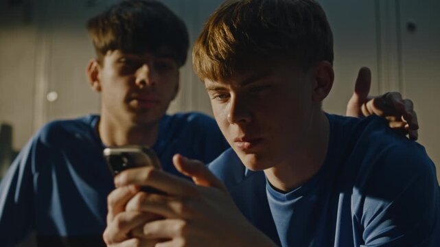 Young Soccer Players in Uniforms in Dressing Room, Sharing Content on Smartphone. One Boy Was Browsing Phone When Teammate Joined For Friendly Chat. Boys Smiling and Discussing Something on Screen.